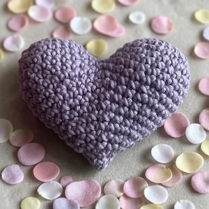 May include: A lavender crocheted heart, a handmade item, rests on a neutral surface, surrounded by scattered pastel-colored flower petals. The heart's texture is visible, showcasing the tight stitches of the crochet work.