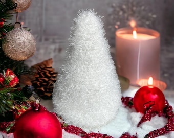 Op de afbeelding: Een witte, donzige kerstboom staat centraal in deze feestelijke scène. Rode en gouden ornamenten, een dennenappel en een brandende kaars dragen bij aan de feestelijke sfeer. Een rode bal en slinger maken de winterse feestdecoratie compleet.