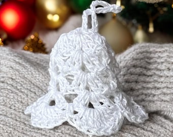 Puede incluir: Adorno de campana de crochet blanco con un lazo en la parte superior. El adorno está sobre una superficie de punto blanco.