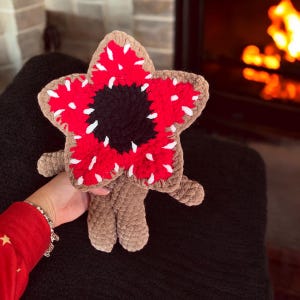 May include: A crocheted Demogorgon plush toy with a distinctive flower-shaped head in red, white, and black, and a brown body. The toy is held up, with a fireplace providing a warm backdrop.
