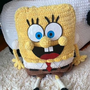 May include: A handmade yellow crochet pillow shaped like the cartoon character SpongeBob SquarePants. The pillow features large white eyes with blue pupils, a wide open mouth, and a red tie. The pillow is on a white surface.