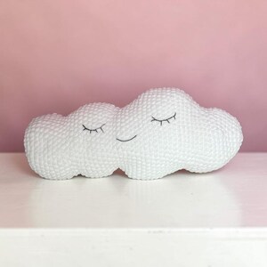 May include: A white cloud-shaped ornament with a textured surface. The cloud features embroidered black closed eyes and a smiling mouth. It rests on a white surface against a pink background.