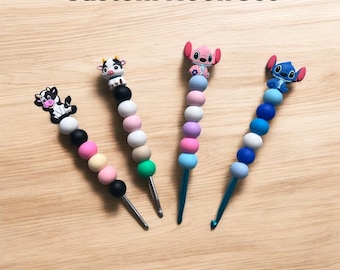 Può includere: Quattro set di uncinetti personalizzati. Ogni uncinetto presenta una serie di perline colorate e un ornamento decorativo. Gli ornamenti includono una mucca, una mucca con un design in bianco e nero e due versioni del personaggio Stitch. Il testo "Custom Hook Set" è in alto.