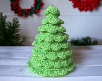 Op de afbeelding: Een groene gehaakte kerstboomversiering met een gegolfd ontwerp. De versiering ligt op een wit oppervlak.