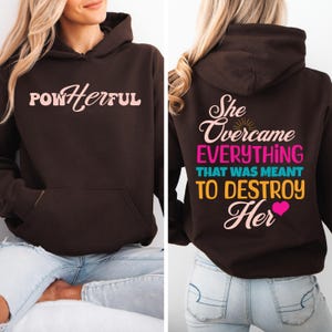 Könnte beinhalten: Brauner Hoodie mit dem Text "POWHerful" auf der Vorderseite und "She Overcame Everything That Was Meant To Destroy Her" auf der Rückseite. Der Text ist in Rosa, Blau, Gelb und Orange.