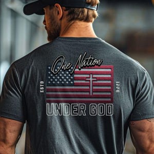 May include: Dark gray t-shirt with a graphic design featuring an American flag with a cross, the words "One Nation Under God," and the year 1776. The shirt has a crew neck and short sleeves.