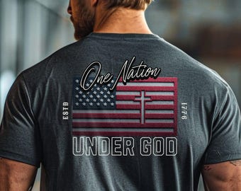 May include: Dark gray t-shirt with a graphic design featuring an American flag with a cross, the words "One Nation Under God," and the year 1776. The shirt has a crew neck and short sleeves.