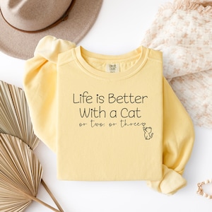 Puede incluir: Sudadera amarilla clara con el texto negro "Life is Better With a Cat or two or three". Una peque&ntilde;a ilustraci&oacute;n de gato est&aacute; al lado. La sudadera est&aacute; doblada sobre una superficie blanca.
