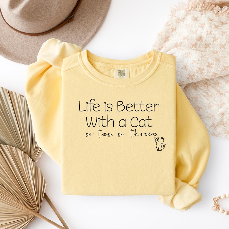 Puede incluir: Sudadera amarilla clara con el texto negro "Life is Better With a Cat or two or three". Una peque&ntilde;a ilustraci&oacute;n de gato est&aacute; al lado. La sudadera est&aacute; doblada sobre una superficie blanca.