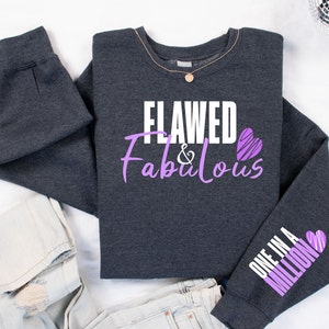 May include: A dark gray sweatshirt with the text "Flawed & Fabulous" in white and purple lettering with a purple heart. The left sleeve has the text "One in a Million" in purple lettering with a purple heart.