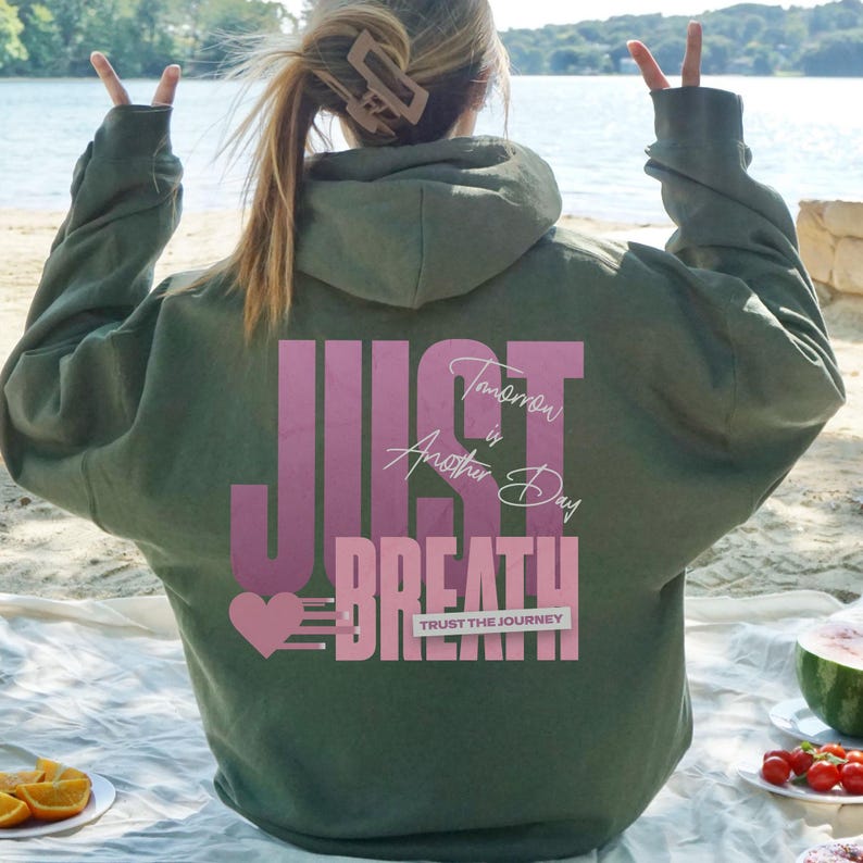 May include: An olive green hoodie with the words "JUST BREATHE" in a gradient pink font. The text "Tomorrow is Another Day" and "TRUST THE JOURNEY" are also printed on the back. A pink heart with lines is on the left side.