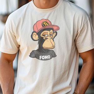 Puede incluir: Una camiseta blanca con una imagen de dibujos animados de un mono con una gorra de béisbol roja con un símbolo de bitcoin. El mono mira hacia la derecha y el texto "FOMO" está impreso debajo de la imagen.