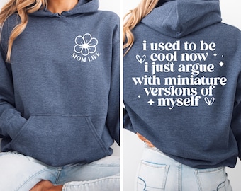 May include: A navy blue hoodie with a white flower graphic on the front and the text "MOM LIFE" inside the flower. The back of the hoodie has the text "i used to be cool now. i just argue with miniature versions of myself" in white.