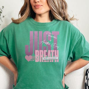 May include: A green t-shirt featuring the words "JUST BREATHE" in a purple gradient font. The text "Tomorrow is Another Day" is above "BREATHE" in white script. A pink heart and "TRUST THE JOURNEY" are also on the shirt.