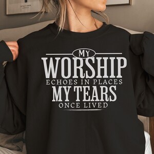 以下が含まれることがあります： 「MY WORSHIP echoes in places MY TEARS once lived」と白い文字がプリントされた黒いスウェットシャツ。テキストは太字のサンセリフ体です。スウェットシャツはクルーネックです。