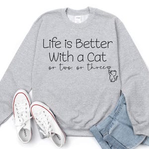 Puede incluir: Una sudadera gris jaspeada con el texto "Life is Better With a Cat or two. or three." en escritura negra. Un peque&ntilde;o dibujo de un gato est&aacute; al lado del texto. Zapatillas blancas y vaqueros azules tambi&eacute;n est&aacute;n en la imagen.