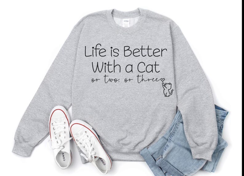 Puede incluir: Una sudadera gris jaspeada con el texto "Life is Better With a Cat or two. or three." en escritura negra. Un peque&ntilde;o dibujo de un gato est&aacute; al lado del texto. Zapatillas blancas y vaqueros azules tambi&eacute;n est&aacute;n en la imagen.