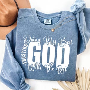 Op de afbeelding: Een blauw sweatshirt met de tekst "Doing My Best Trusting God With The Rest" in witte letters.