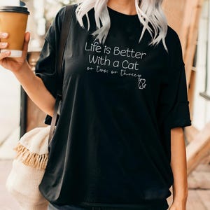 Puede incluir: Camiseta negra con la frase blanca "Life is Better With a Cat or two or three" y un dibujo de un gato. La camiseta es de estilo casual.