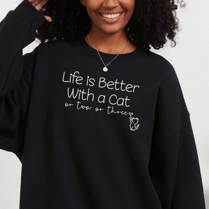 Puede incluir: Sudadera negra con el texto blanco "Life is Better With a Cat or two or three". Una peque&ntilde;a ilustraci&oacute;n de gato blanco acompa&ntilde;a el texto. La sudadera es de cuello redondo.