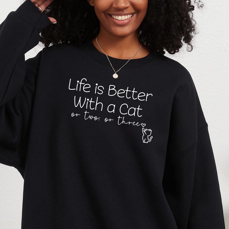 Puede incluir: Sudadera negra con el texto blanco "Life is Better With a Cat or two or three". Una peque&ntilde;a ilustraci&oacute;n de gato blanco acompa&ntilde;a el texto. La sudadera es de cuello redondo.