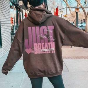May include: A brown hoodie featuring the words "JUST BREATHE" in a purple and pink design on the back. The text "Tomorrow is Another Day" is above the word "BREATHE". The phrase "TRUST THE JOURNEY" is below the word "BREATHE".