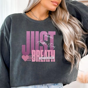 May include: Dark grey sweatshirt featuring the words "JUST BREATHE" in a pink gradient font. The text "Tomorrow is Another Day" is above the word "BREATHE". A pink heart and the words "TRUST THE JOURNEY" are also visible.