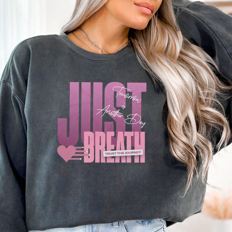 May include: Dark grey sweatshirt featuring the words "JUST BREATHE" in a pink gradient font. The text "Tomorrow is Another Day" is above the word "BREATHE". A pink heart and the words "TRUST THE JOURNEY" are also visible.