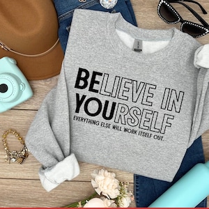 May include: A light gray sweatshirt with the text "BELIEVE IN YOURSELF" in black. The sweatshirt is paired with blue jeans, a brown hat, sunglasses, a turquoise camera, a bracelet, and a turquoise water bottle.