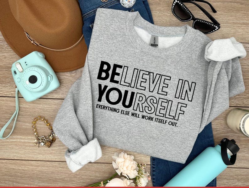 May include: A light gray sweatshirt with the text "BELIEVE IN YOURSELF" in black. The sweatshirt is paired with blue jeans, a brown hat, sunglasses, a turquoise camera, a bracelet, and a turquoise water bottle.