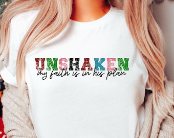 May include: White t-shirt with the text "UNSHAKEN my faith is in his plan" in colorful, distressed letters.