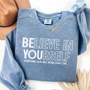 May include: A blue sweatshirt with the words "BELIEVE IN YOURSELF" printed in white. The phrase "EVERYTHING ELSE WILL WORK ITSELF OUT" is printed below. A gold necklace and a brown hat are also visible.