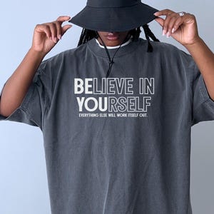 May include: A dark gray t-shirt with the words "BELIEVE IN YOURSELF" in white block letters. Below the text is the phrase "EVERYTHING ELSE WILL WORK ITSELF OUT." A black bucket hat is worn.