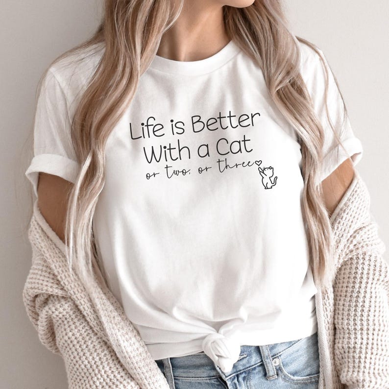 Puede incluir: Camiseta blanca con el texto negro "Life is Better With a Cat or two or three". Una peque&ntilde;a ilustraci&oacute;n de gato negro acompa&ntilde;a el texto. Estilo casual.