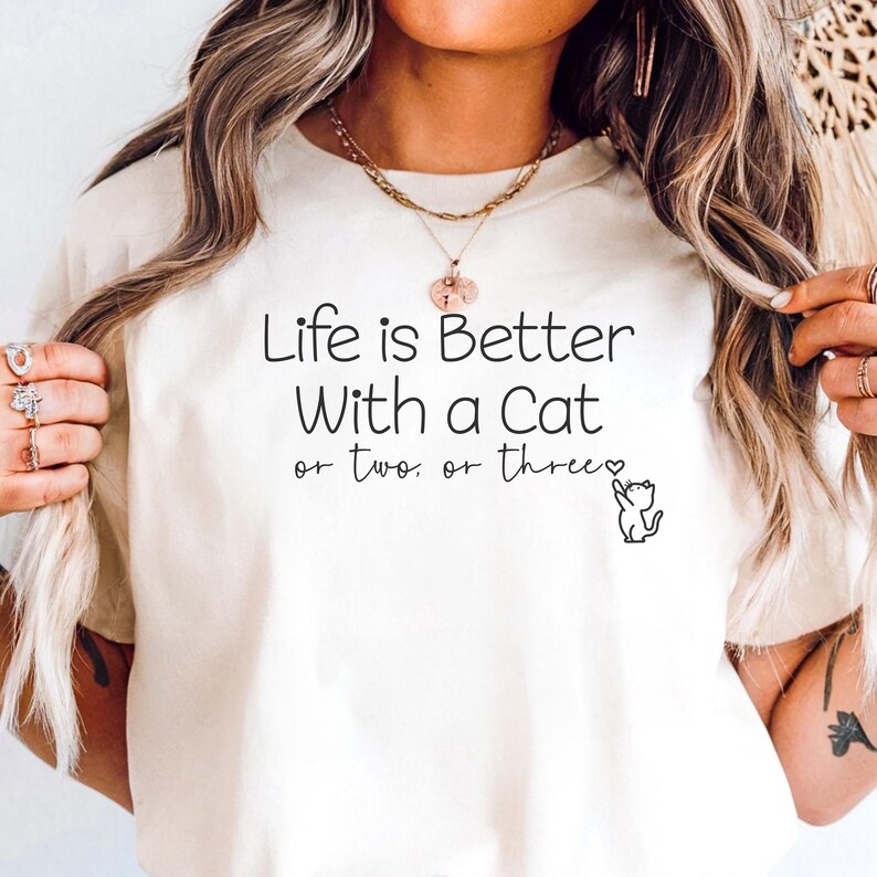 Puede incluir: Camiseta color crema con el texto negro "Life is Better With a Cat or two or three". Una peque&ntilde;a ilustraci&oacute;n de gato negro acompa&ntilde;a el texto. La camiseta es de estilo casual.