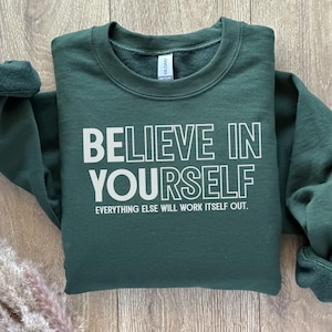 May include: A dark green sweatshirt with the words "BELIEVE IN YOURSELF" in white block letters. Below the main text is the phrase "EVERYTHING ELSE WILL WORK ITSELF OUT." The sweatshirt has a crew neck and long sleeves.