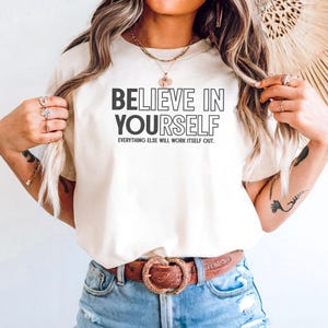 May include: Cream-colored t-shirt with the text "BELIEVE IN YOURSELF" in a bold, black font. Below the main text, the phrase "EVERYTHING ELSE WILL WORK ITSELF OUT" is printed in a smaller, black font. The shirt is paired with blue denim shorts and a brown belt.