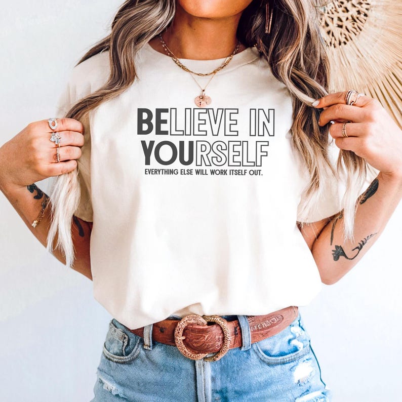 May include: Cream-colored t-shirt with the text "BELIEVE IN YOURSELF" in a bold, black font. Below the main text, the phrase "EVERYTHING ELSE WILL WORK ITSELF OUT" is printed in a smaller, black font. The shirt is paired with blue denim shorts and a brown belt.