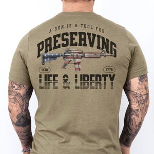 May include: A green t-shirt with the text "A gun is a tool for preserving life & liberty" and a stylized image of a rifle with an American flag design. The text "Estd 1776" is also included.