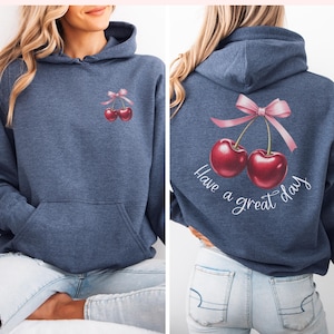 May include: A navy blue hoodie with a pocket on the front and a graphic on the back. The graphic features two red cherries with a pink bow and the text "Have a great day".