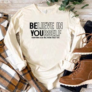 May include: Cream-colored long-sleeve shirt with the text "BELIEVE IN YOURSELF" in black. The phrase "EVERYTHING ELSE WILL WORK ITSELF OUT" is below. The shirt is paired with a brown plaid shirt, dark jeans, and brown boots.