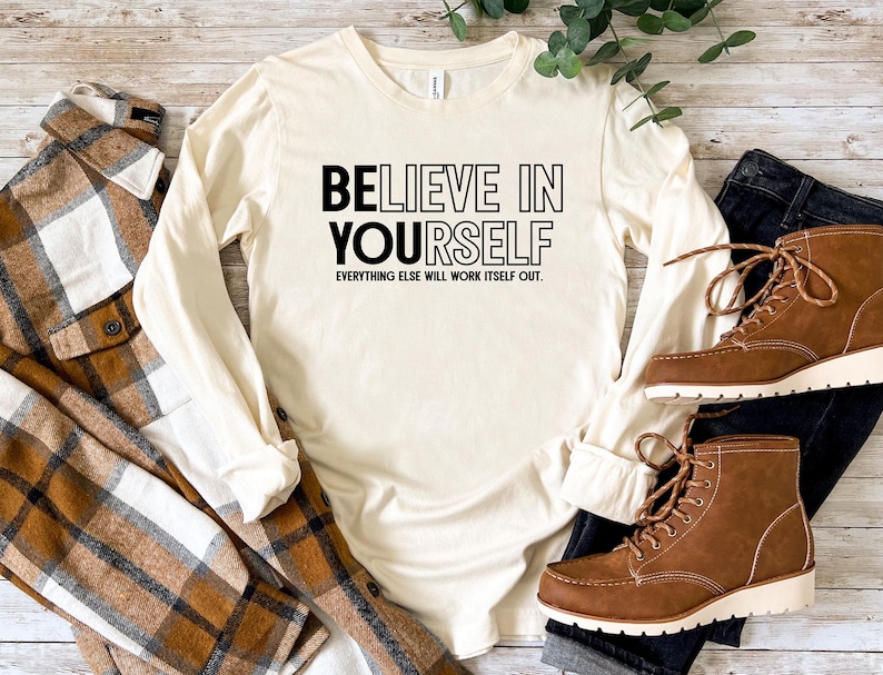 May include: Cream-colored long-sleeve shirt with the text "BELIEVE IN YOURSELF" in black. The phrase "EVERYTHING ELSE WILL WORK ITSELF OUT" is below. The shirt is paired with a brown plaid shirt, dark jeans, and brown boots.