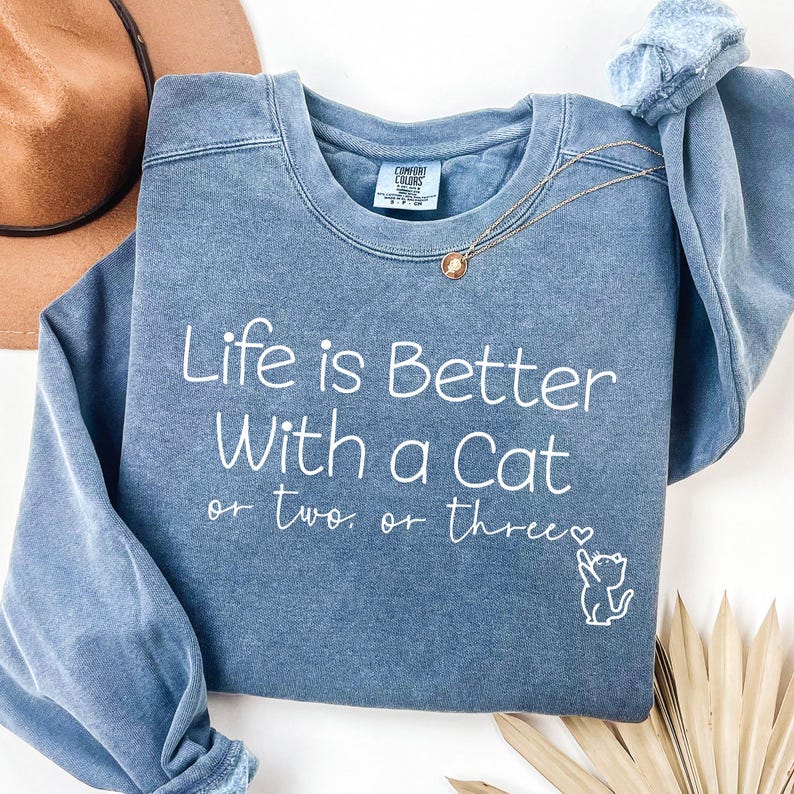 Puede incluir: Sudadera azul con la frase blanca "Life is Better With a Cat or two or three" y un peque&ntilde;o gr&aacute;fico de gato. Un collar dorado est&aacute; sobre la sudadera.