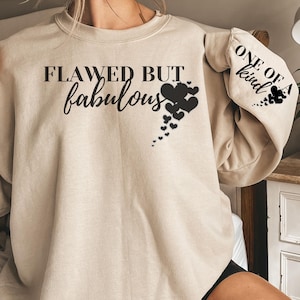 Op de afbeelding: Een beige sweatshirt met de tekst "Flawed But Fabulous" en een ontwerp van zwarte harten op de voorkant. De linkermouw heeft de tekst "One of a kind" in zwart.
