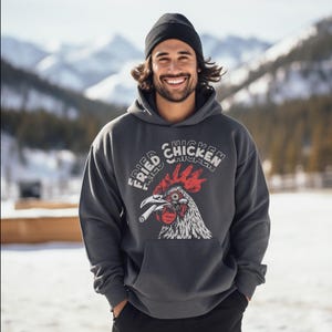 Peut inclure: Un sweat-shirt gris foncé avec une poche avant et un motif de coq fumant une cigarette, avec l'inscription "FRIED CHICKEN" en arc de cercle. La personne porte un bonnet noir.