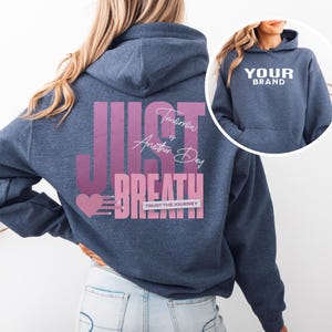 May include: A navy blue hoodie featuring the words "JUST BREATHE" in a pink and white design. The text "Tomorrow is Another Day" is also printed on the back. The hoodie has a front pocket and a hood.
