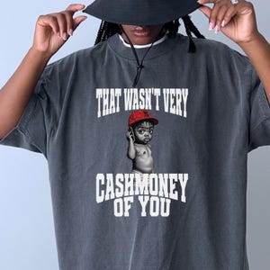Op de afbeelding: Donkergrijs t-shirt met de tekst "THAT WASN'T VERY" boven een illustratie van een baby met een rode pet en een telefoon. De tekst "CASHMONEY OF YOU" staat eronder. De persoon draagt een zwarte hoed.