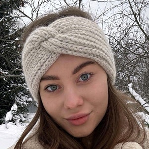 May include: A woman wearing a white knitted headband and a white faux fur coat. She is standing in a snowy outdoor setting.