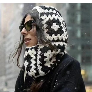 Op de afbeelding: Een zwart-witte gebreide capuchon sjaal met een geometrisch patroon. De sjaal wordt gedragen door een persoon met lang donker haar en een zonnebril.