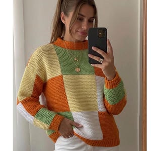 May include: A long-sleeve sweater featuring a color-blocked patchwork design in shades of orange, yellow, white, and light green. The garment has a high neckline and a textured knit pattern throughout, with ribbed cuffs and a ribbed hemline.